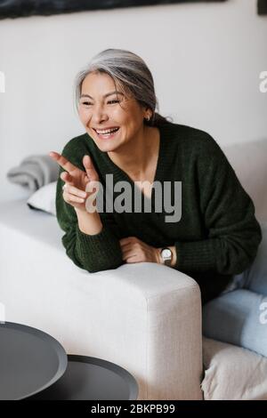 Foto von grauhaarigen freudigen Frau, die mit dem Finger zur Seite zeigt und lacht, während sie im Wohnzimmer auf der Couch sitzt Stockfoto