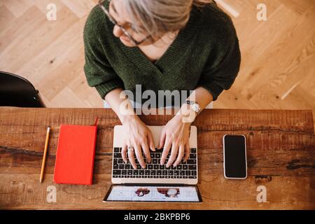 Foto von silbernen reifen Frau in Brillen mit Laptop während am Tisch im Wohnzimmer sitzen Stockfoto