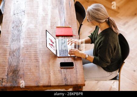 Foto von silbernen reifen Frau in Brillen mit Laptop während am Tisch im Wohnzimmer sitzen Stockfoto
