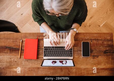 Foto von silbernen reifen Frau in Brillen mit Laptop während am Tisch im Wohnzimmer sitzen Stockfoto