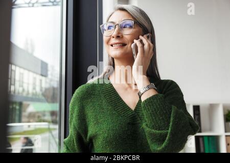 Foto von grauhaarigen lächelnden Geschäftsfrau in Brillen sprechen auf Handy während der Arbeit im Büro Stockfoto