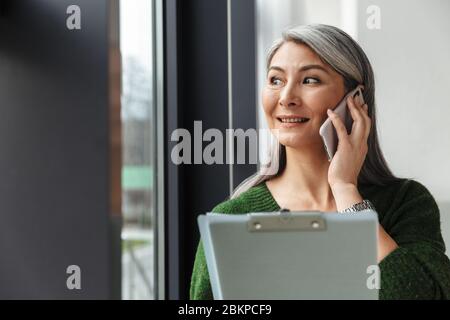 Foto von grauhaariger lächelnder Geschäftsfrau, die während der Arbeit mit Dokumenten im Büro auf dem Handy spricht Stockfoto