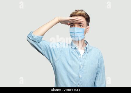 Porträt von aufmerksamen schönen jungen Mann mit chirurgischen medizinischen Maske in blauem Hemd mit der Hand auf der Stirn stehen und mit ernsten Gesicht wegschauen. ich Stockfoto