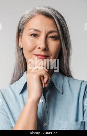 Bild von erwachsenen reifen Frau mit langen weißen Haaren tragen Hemd berühren Kinn und Blick auf Kamera isoliert über grauem Hintergrund Stockfoto