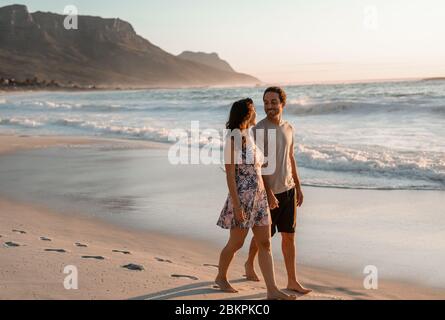 Liebende junge Paare, die an einem Sandstrand entlang spazieren Stockfoto