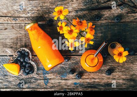 Orange und Karotten Smoothie in Glasflaschen mit rostfreiem Stroh und Kristall kleine Vasen mit Chernobrivtsi Blumen und Heidelbeeren auf rustikalem Grunge Holzhintergrund. Stockfoto