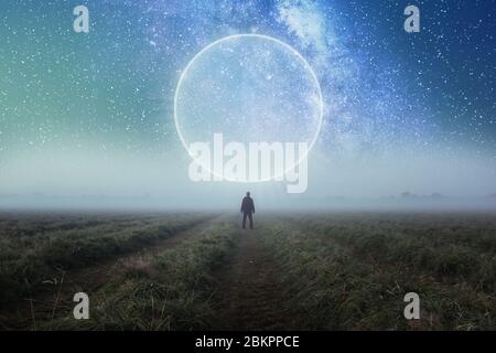 Ein Science-Fiction-Konzept. Ein Mann, der in einem Feld steht und über den Raum blickt, mit einem glühenden Portal am Nachthimmel Stockfoto