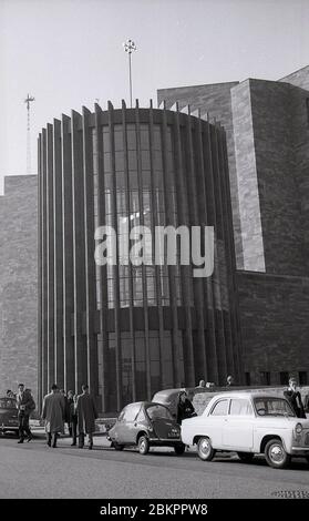 1961, historisches Bild, das Menschen vor dem Eingang der neuen Kathedrale, Coventry, England bei ihrer Eröffnung zeigt, mit Autos der Zeit auf der Straße geparkt, darunter ein 'Bubble Car' und ein Austin. Die Stadt wurde während des Blitzes im 2. Weltkrieg schwer bombardiert und musste in den späteren Jahrzehnten wieder aufgebaut werden, als an der Stelle der Ruinen der alten eine neue "moderne" Kathedrale errichtet wurde. Das neue Gebäude wurde von Basil Spence entworfen, einem Architekten, der für seine radikalen 'Brutalistischen' Strukturen berühmt ist. Stockfoto