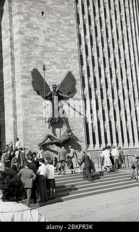 1961, historisches Bild von Menschen vor der neuen Kathedrale, Coventry, England bei ihrer Eröffnung. Die Skulptur an der Wand von Jacob Epstein bezeichnet den Sieg des hl. Michaels über den Teufel. Im Mai 1962 wurde ein neues "modernistisches" Gebäude offiziell auf dem Gelände der Ruinen der alten Kathedrale eröffnet. Das Gebäude wurde von Basil Spence im radikalen 'Brutalist'-Architekturstil entworfen, der in dieser Zeit üblich ist. Stockfoto