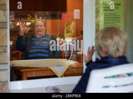 Balve, Deutschland. Mai 2020. Engelbert Prumbaum winkt seiner Tochter Andrea Rapp aus dem Fenster des St. Johannes-Altenheim. Während er hinter einer Plexiglasscheibe in der ungenutzten Cafeteria der Anlage sitzt, hat sie sich draußen auf einem Stuhl gesetzt. Alte Menschen- und Pflegeheime in NRW tun viel, um auch bei der Corona-Pandemie Verwandten einen Besuch zu ermöglichen. Quelle: Bernd Thissen/dpa/Alamy Live News Stockfoto