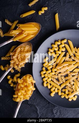 Drei Besteck Löffel und Teller mit Vielzahl von roh goldenen Weizen Pasta auf dunkelschwarz strukturierten Hintergrund, Draufsicht Stockfoto