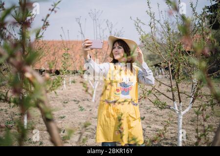 Frau, die ihren Sonnenhut berührt, während sie im Hof steht. Porträt von Mädchen posiert während der Aufnahme Selfie im Garten mit verschwommenen Zweigen und Knospen. Stockfoto