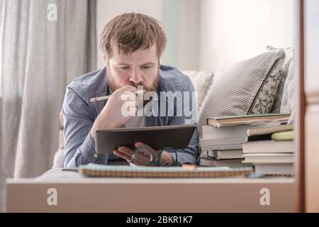 Junger nachdenklicher Mann, der fernab studiert, während er mit einem Tablet und Büchern auf einem Sofa liegt. Konzept der Online-Bildung während einer Quarantäne. Stockfoto