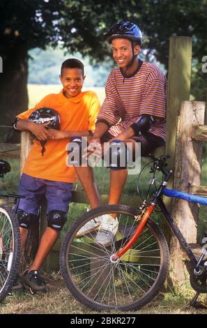 Fahrradfahrräder Teenage Britisch-afrikanisch-karibisch-amerikanische Brüder 11 Jahre -15 Jahre mit Helmen und schützendem Fahrradkit, draußen in der Sommersonne, die während ihrer Fahrradabenteuer während der Sommerschulferien eine Pause einlegen. Stockfoto