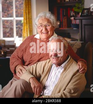 Ältere Paar Senioren Rentner glücklich gesundes Paar 80-86 Jahre Hausbesitzer sitzen innen in ihrem komfortablen Wohnzimmer Gegenüberliegende Kamera Stockfoto