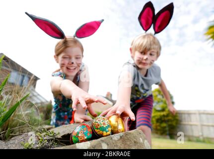 Ein Bruder und eine Schwester, die Gesichtsfarbe und hausgemachte Hasenohren tragen, nehmen an einer Ostereiersuche am Ostersonntag in ihrem Garten Teil. Stockfoto