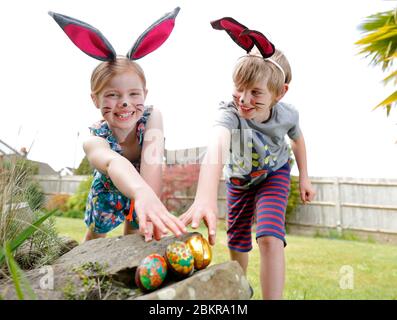 Ein Bruder und eine Schwester, die Gesichtsfarbe und hausgemachte Hasenohren tragen, nehmen an einer Ostereiersuche am Ostersonntag in ihrem Garten Teil. Stockfoto