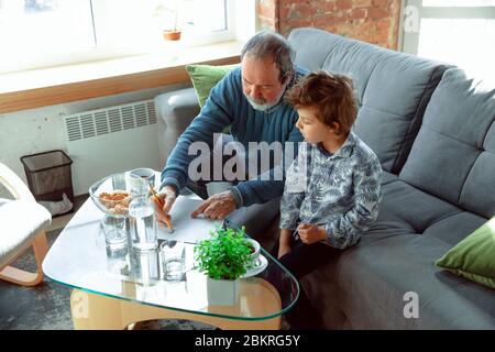 Nützliche Zeit. Großvater und sein Enkel verbringen Zeit zusammen isoliert zu Hause. Spaß haben, Stadtmachen, Lesen und Zeichnen. Konzept der Quarantäne, Familie, Liebe, Realismus, Zweisamkeit, Bildung. Stockfoto