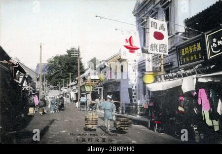 [ Japan des 19. Jahrhunderts - Japanische Geschäfte in Yokohama ] - EIN Verkäufer, der seine Waren trägt, geht an Geschäften in Motomachi, Yokohama, Kanagawa Präfektur vorbei. Vintage-Postkarte des 20. Jahrhunderts. Stockfoto