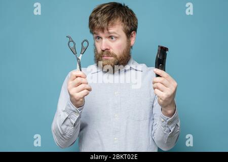 Rätselhafter, zotteliger Mann mit Schere und Trimmer in den Händen entscheidet, womit er sich die Haare schneiden soll. Konzept der Quarantäne während einer Pandemie. Stockfoto