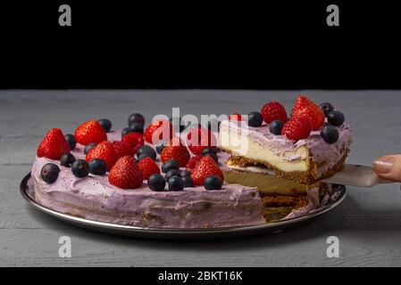 Frau geteilt Käsekuchen und nehmen Sie eine Scheibe, um es anzubieten. Hausgemachtes Käsekuchen mit Erdbeere, Heidelbeere und Himbeere auf grauem Holzhintergrund. Stockfoto