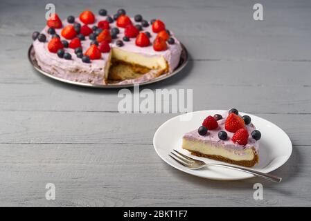 Scheibe hausgemachter Käsekuchen mit Beeren auf dem Teller. Käsekuchen mit Erdbeere, Heidelbeere und Himbeere auf grauem Holzhintergrund. Stockfoto