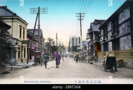 [ Japan des 19. Jahrhunderts - Japanische Geschäfte in Yokohama ] - die Menschen kommen an Geschäften auf Otamachi-dori (太田町道り) in Yokohama, Präfektur Kanagawa vorbei. Auf dem Schild auf dem gelben Gebäude steht Daikokuya, daneben befindet sich der Sonnenschirmladen Isekan. Karren und Rikschas werden entlang der Bürgersteige geparkt. Vintage-Postkarte des 20. Jahrhunderts. Stockfoto