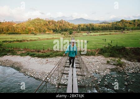 Malaysia, 3. Mai 2020 - Reisbauer überquert Hängebrücke nach der Arbeit mit Reisfeld im Hintergrund im Tegudon Tourism Village in Kota Belud, Stockfoto