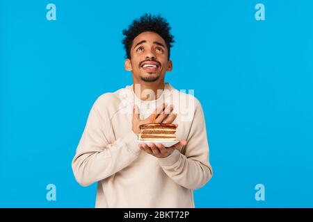 Aufrichtig und hoffnungsvoll afrikanisch-amerikanischer Kerl, der den b-Tageskuchen mit Kerze hält, Hand am Herzen hält und den Himmel schaut, während er den Wunsch erfüllt Stockfoto