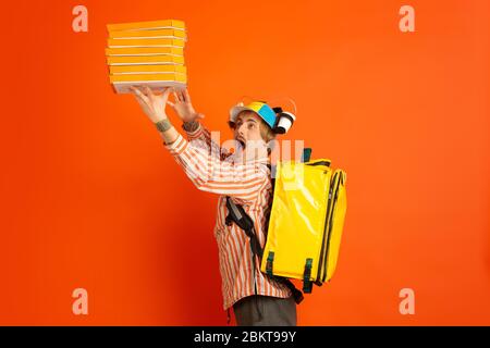 Anmutige Pizzaschachteln. Emotionen des kaukasischen Deliveryman isoliert auf orangefarbenem Hintergrund. Kontaktloser Lieferservice während der Quarantäne. Der Mensch liefert Nahrung während der Isolation. Sicherheit. Schnell nach oben. Stockfoto