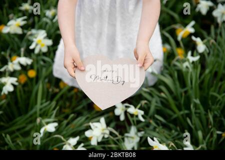 Happy Mother's Day auf einer Papierkarte geschrieben, die von einem Mädchen auf einem Blumen- und Grashintergrund gehalten wird Stockfoto