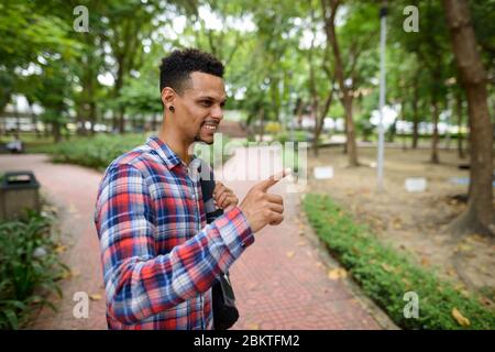 Fröhlicher junger afrikanischer Hipster-Mann mit Rucksack, der mit dem Finger auf den Park zeigt Stockfoto