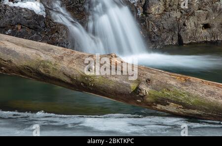 Ein Holzklöster liegt auf einer dünnen Eisschicht auf einem Fluss in Arkansas mit einem Wasserfall im Hintergrund. Stockfoto