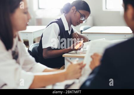 Schüler in Uniform sitzen am Schreibtisch im Klassenzimmer. Studentin im Klassenzimmer mit Klassenkameradin, die vor der Tür diskutiert. Stockfoto