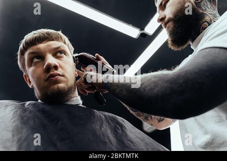 Männlicher Haarschnitt mit Rasiermesser. Tätowierte Barber macht Haarschnitt für Kunden im Friseur-Shop mit Haarschneidemaschine. Mann Friseursalon mit Elektro Stockfoto