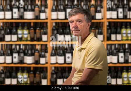 Älterer alter Mann, der die Kamera mit einem Hintergrund eines Weinkellers voller Tausende von Flaschen in Regalen betrachtet Stockfoto