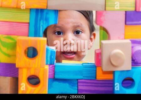 Ein kleiner Junge zeigt sein Gesicht durch ein Fenster hinter einer Wand aus Bausteinen mit verschiedenen Formen und Farben. Stockfoto