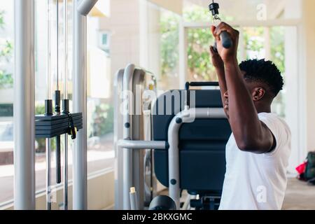 Starker afroamerikanischer Mann in Sportswear, der während des Trainings im Fitnessstudio Kabel mit Bar zieht und auf dem Trainingsgerät sitzt. Stockfoto