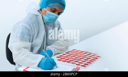 Arzt mit Maske und blauen Handschuhen, die auf einem weißen Blatt Papier schreiben, das neben einer Gruppe von klinischen Proben roter Flüssigkeit in runden weißen Behältern sitzt Stockfoto