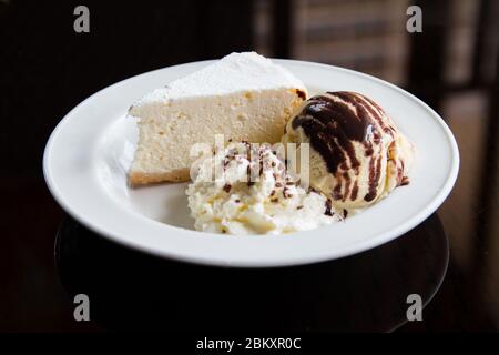 Eine Scheibe Käsekuchen mit Eis und Schlagsahne auf einem weißen Teller. Konzeptuelle Food-Fotografie mit Pflanzendekoration. Stockfoto