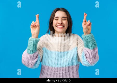 Optimistische, niedliche junge Frau im Winter-Pullover, die Glaubensträume haben, werden wahr, lächelnd breit, nahe Augen, die sich mit den Daumen wünschen Stockfoto