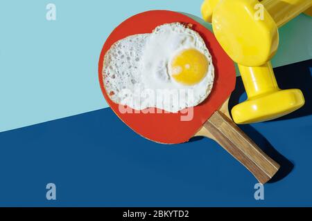 Auf dem blauen Tisch sind gelbe Hanteln, ein roter Tischtennisschläger und Eier. Eier mit einem dünnen Eigelb liegen auf Rucola. Stockfoto