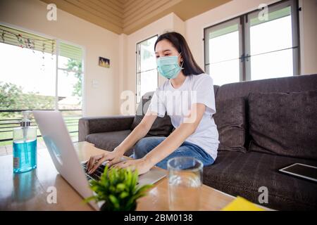 Asiatische Frau niedlich schön trägt Schutzmaske und mit einem Notebook-Computer und sitzt auf dem Sofa für die Arbeit von zu Hause aus. Sie ist bei h unter Quarantäne gestellt Stockfoto