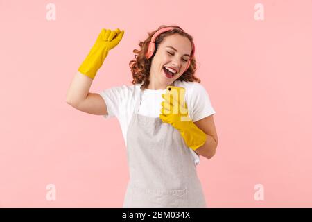 Foto einer glücklichen singenden jungen Frau Hausfrau in Handschuhen isoliert über rosa Wand Hintergrund mit Handy Musik hören mit Kopfhörern. Stockfoto