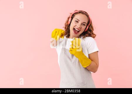 Foto einer glücklichen singenden jungen Frau Hausfrau in Handschuhen isoliert über rosa Wand Hintergrund mit Handy Musik hören mit Kopfhörer pointin Stockfoto