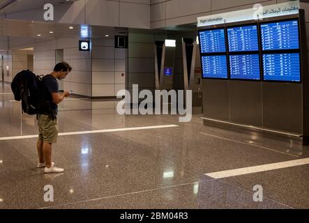 Ein westlicher Backpacker überprüft sein Smartphone und seine Abflugtafeln am internationalen Flughafen Hamad in Doha, Katar. Stockfoto