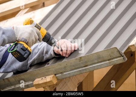 Männliche Hände in Arbeitshandschuhe mit einem gelben Schraubendreher Schrauben die Dachplatte auf das Dach des Landhauses. Akku-Bohrmaschine. Stockfoto