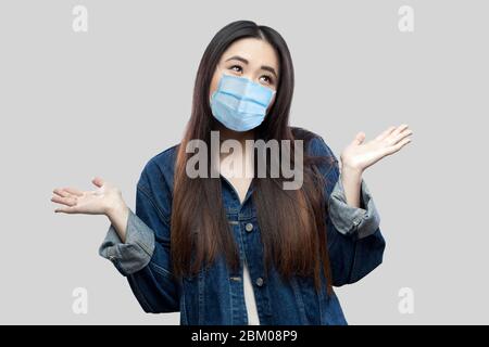 Ich weiß es nicht. Verwirrt schöne brünette asiatische junge Frau mit chirurgischen medizinischen Maske in blauen Denim Jacke stehend erhobenen Armen und wegschauen Thou Stockfoto