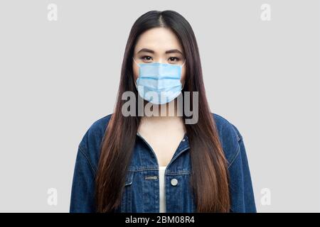 Porträt der schönen Brünette asiatische junge Frau mit chirurgischen medizinischen Maske in blauen Denim Jacke stehen und Blick auf die Kamera. Indoor-Studio geschossen, ich Stockfoto