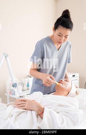 Asiatische Kosmetikerin in Peelings Anwendung Gesichtsmaske auf reife Kunden Gesicht mit Pinsel im Schönheitssalon Stockfoto
